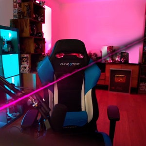 Op de afbeelding: Een zwarte en blauwe gamingstoel met witte accenten en het woord "DXRACER" gedrukt op de hoofdsteun. De stoel staat in een kamer met roze en blauwe verlichting en planken vol met verschillende items.