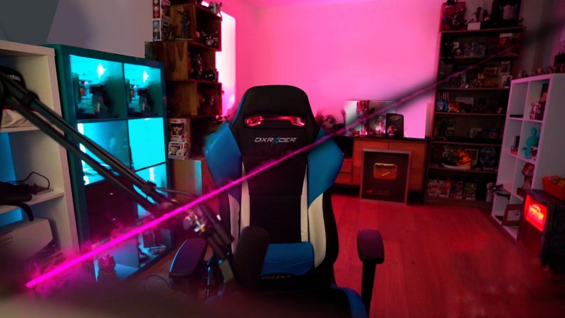 Op de afbeelding: Een zwarte en blauwe gamingstoel met witte accenten en het woord "DXRACER" gedrukt op de hoofdsteun. De stoel staat in een kamer met roze en blauwe verlichting en planken vol met verschillende items.