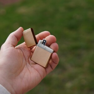 May include: A gold-colored lighter with a silver insert is open and held in a hand. The lighter is against a green grassy background.