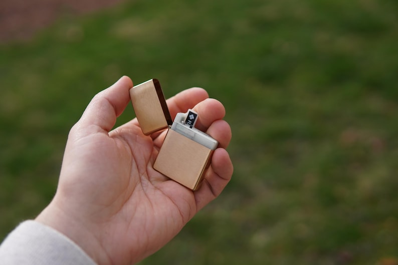 May include: A gold-colored lighter with a silver insert is open and held in a hand. The lighter is against a green grassy background.