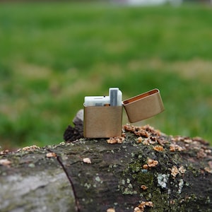 May include: A gold metal lighter with a rectangular shape and a hinged lid. The lighter is open and sitting on a brown log.