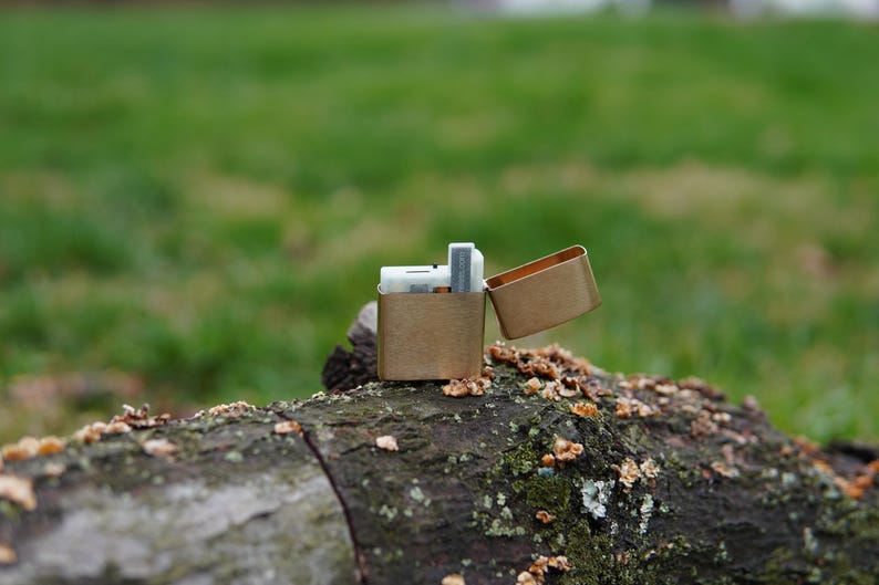 May include: A gold metal lighter with a rectangular shape and a hinged lid. The lighter is open and sitting on a brown log.