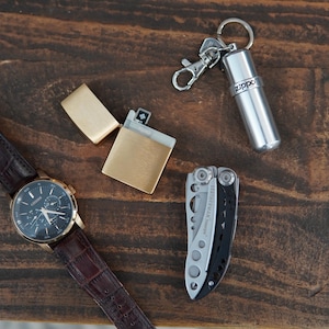 May include: A brown leather wristwatch with a black face and silver hands, a gold lighter, a silver multi-tool, and a silver keychain with a small silver container with the word "Zippo" printed on it.
