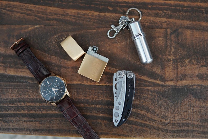 May include: A brown leather wristwatch with a black face and silver hands, a gold lighter, a silver multi-tool, and a silver keychain with a small silver container with the word "Zippo" printed on it.