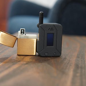 May include: A gold lighter with a black and silver device next to it. The device has a small screen and a button.