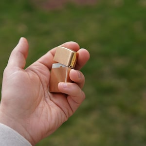 May include: A gold-colored lighter with a white interior is held in a person's hand. The lighter is closed and appears to be made of metal.