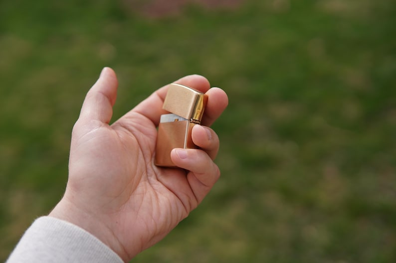 May include: A gold-colored lighter with a white interior is held in a person's hand. The lighter is closed and appears to be made of metal.