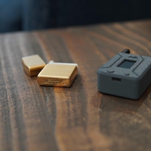 May include: Three small rectangular objects on a wooden surface. Two are gold-colored metal lighters and one is a black electronic device with a small screen.