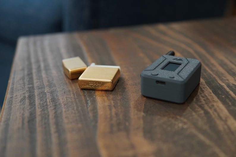 May include: Three small rectangular objects on a wooden surface. Two are gold-colored metal lighters and one is a black electronic device with a small screen.