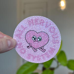 Puede incluir: Una pegatina redonda rosa con un corazón de dibujos animados triste y el texto "ALL NERVOUS SYSTEM". El corazón tiene brazos y piernas. La pegatina tiene un efecto brillante.