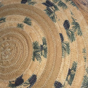 May include: A circular placemat with a spiral design. The placemat is made of tan fabric with a repeating pattern of green pine needles and pine cones. The design is stitched with white thread.
