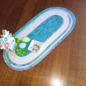 May include: An oval-shaped table runner with a teal and white design. The runner is decorated with a small floral arrangement, two pastel-colored eggs, and a small square dish with a botanical design. The runner is on a wooden table.