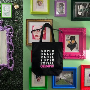 May include: A black tote bag with white text that reads "SUPER CALIF RAGIL ISTIC EXPIAL OZEMPIC". The bag is hanging on a wall with several framed pictures and a mirror.