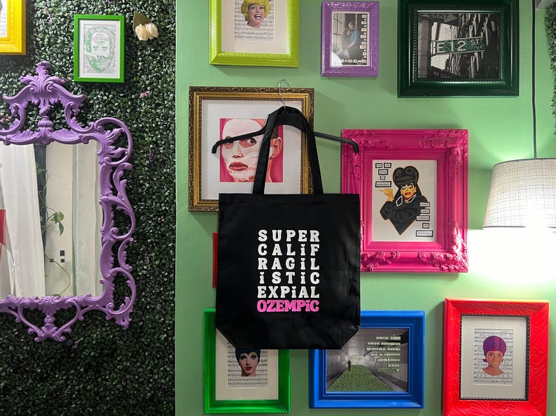 May include: A black tote bag with white text that reads "SUPER CALIF RAGIL ISTIC EXPIAL OZEMPIC". The bag is hanging on a wall with several framed pictures and a mirror.