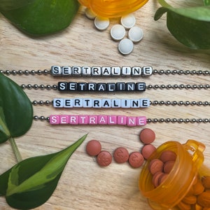 Puede incluir: Cuatro cadenas plateadas con la palabra "SERTRALINE" escrita en cuentas de letras negras, blancas y rosas. Las cadenas están sobre una superficie de madera con una planta verde, un frasco de pastillas y pastillas blancas.