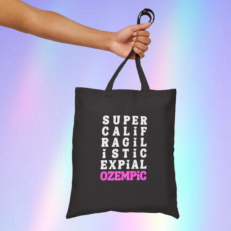 May include: Black tote bag with white text that reads "SUPER CALIF RAGIL ISTIC EXPIAL OZEMPIC" with the word "OZEMPIC" in pink.