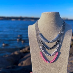 Puede incluir: Un collar de capas con el nombre "Jennifer Coolidge" escrito en cuentas blancas sobre una cadena plateada. El collar se muestra en un maniquí sobre un fondo de agua azul.