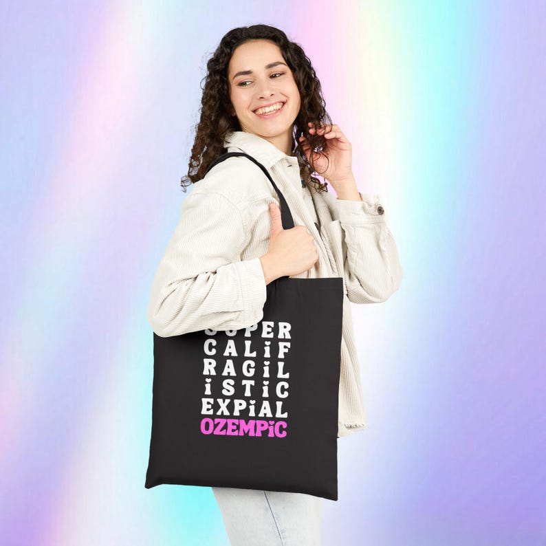 May include: A black tote bag with white text that reads "SUPER CALIF RAGIL ISTIC EXPIAL OZEMPIC" in a grid pattern. The text "OZEMPIC" is in pink.