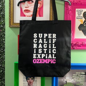 May include: Black canvas tote bag with white text that reads "SUPER CALI RAGIL ISTIC EXPIAL OZEMPIC" with the word "OZEMPIC" in pink.