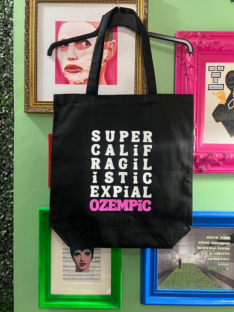 May include: Black canvas tote bag with white text that reads "SUPER CALI RAGIL ISTIC EXPIAL OZEMPIC" with the word "OZEMPIC" in pink.