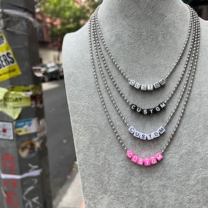Op de afbeelding: Vier zilveren kettingen met het woord "CUSTOM" gespeld in zwarte, witte en roze kralen.