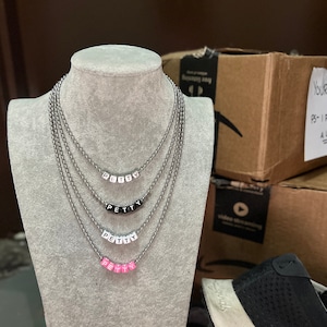 Puede incluir: Un collar de cadena plateada con cuatro capas. Cada capa tiene la palabra "PETTY" escrita en cuentas de letras negras, blancas y rosas.