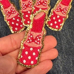 May include: Four pink and red lipstick-shaped iron-on patches with gold glitter trim and white polka dots. Each patch has a pink bow on the top.