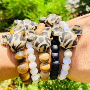 May include: A stack of bracelets with different patterns and colors. The bracelets include brown wood beads, white beads, black beads, and leopard print silicone beads.