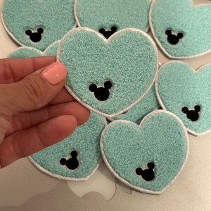 May include: Six turquoise heart-shaped patches with black Mickey Mouse ears in the center. The patches have a white border.