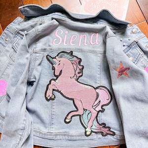 Puede incluir: Una chaqueta vaquera azul claro con un parche de unicornio rosa, un parche de corazón rosa, un parche de estrella y el nombre "Siena" bordado en la espalda.