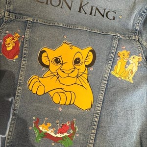 May include: A blue denim jacket featuring The Lion King graphics. The back displays a large Simba illustration, smaller character decals, and the text LION KING. The design is accented with scattered clear rhinestones across the fabric surface.