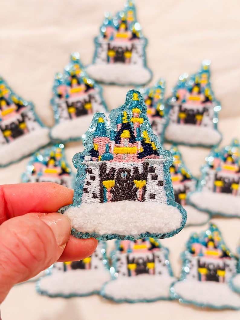 K&ouml;nnte beinhalten: Ein Nahaufnahme eines blauen und rosa Glitzer-Schloss-Patches mit einer wei&szlig;en Wolkenbasis. Der Patch ist mit Details bestickt und hat eine glitzernde Oberfl&auml;che.