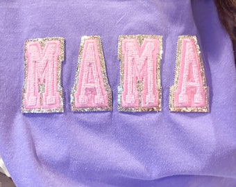 May include: A lilac-colored sweatshirt with the word "MAMA" appliquéd across the chest. The letters are pink with gold glitter borders. The sweatshirt is made of a soft, comfortable material.