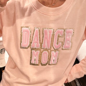 Könnte beinhalten: Ein rosa Sweatshirt mit dem Schriftzug "Dance Mom" in goldenen und rosafarbenen Glitzerbuchstaben.