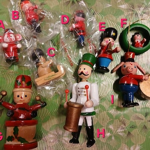 May include: A collection of vintage Christmas ornaments, including Santa figures, a soldier, a pig playing a drum, and a figure in a green wreath. The ornaments are made of wood and painted in red, white, and blue. Some are still in their original packaging.