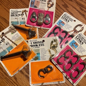 May include: A collection of six packages containing various metal hooks and brackets for hanging planters. The packages are labeled with the brand name "Homescraft" and descriptions of the products, such as "Universal Mount Bracket", "Decorative Connecting Links", and "#1 Swivel Hook/Eye".