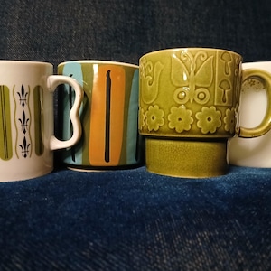 Könnte beinhalten: Vier Vintage-Keramikbecher mit einzigartigen Designs. Ein Becher ist weiß mit grünen und schwarzen vertikalen Akzenten. Ein anderer hat blaue, orange und schwarze Streifen. Ein grüner Becher zeigt Blumen- und Schmetterlingsmuster. Der letzte Becher ist weiß mit einem Blumenmuster.
