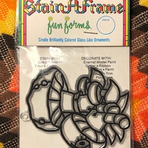 May include: A black and white outline of a cartoon character with a large eye and a hat. The character is on a white card with the text "Stain-A-Frame fun forms Create Brilliantly Colored Glass-Like Ornaments STAIN WITH: Great Glass TM Titan & D Skins DECORATE WITH: Enamel Model Paint Ger Ribbon Gutter Paint Bea Po Trim"