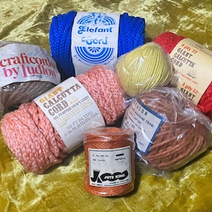 May include: A collection of craft cords and twine in various colors, including blue, red, peach, yellow, brown, and orange. The image shows several spools and bundles of different sizes and textures, with labels indicating the type and length of the cord.