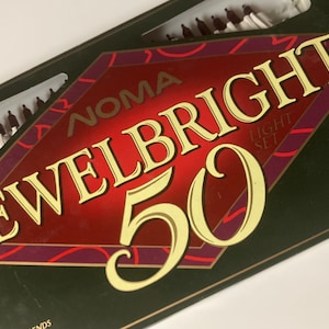Puede incluir: Una caja de luces NOMA Jewelbright 50. La caja es de color verde oscuro con un diseño en forma de diamante rojo y dorado. El texto "JEWELBRIGHT 50" está en letras doradas. La caja contiene pequeñas luces blancas.