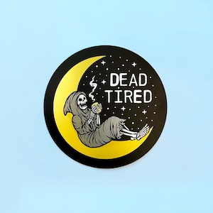 May include: A black and gold sticker with a skeleton sitting on a crescent moon. The skeleton is holding a cup of coffee and the text "DEAD TIRED" is above it.