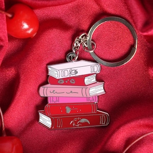 Puede incluir: Un llavero con una pila de libros en tonos rosas y rojos, con detalles metálicos plateados. Los libros tienen páginas blancas y detalles decorativos. El llavero está sobre un fondo de satén rojo con cerezas.