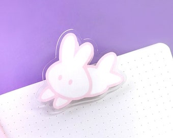 Puede incluir: Un marcapáginas de plástico transparente con un diseño de conejo rosa. El conejo está tumbado de lado con la cabeza girada hacia un lado. El marcapáginas está en una libreta blanca con puntos.