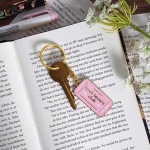 May include: A gold key with a pink "Book Lovers Club" tag on a gold ring rests on an open book. The tag has the text "Admit One" and "Lifetime Membership". A pen and flowers are also visible.