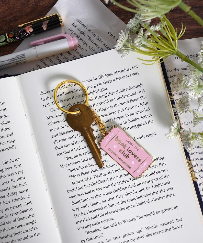 May include: A gold key with a pink "Book Lovers Club" tag on a gold ring rests on an open book. The tag has the text "Admit One" and "Lifetime Membership". A pen and flowers are also visible.