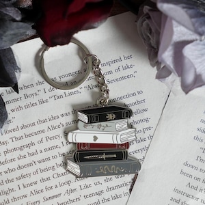 Może przedstawiać: Srebrny brelok do kluczy z zawieszką przedstawiającą stos książek. Książki są w odcieniach czerni, szarości, bieli i bordo, ze srebrnymi detalami. Brelok jest przymocowany do srebrnego kółka i łańcuszka. Książki mają około 5 cm wysokości.