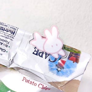 Op de afbeelding: Een transparante plastic chipclip met een roze konijnenontwerp. Het konijn is wit met roze oren en een roze omtrek. De clip is bevestigd aan een zak chips.