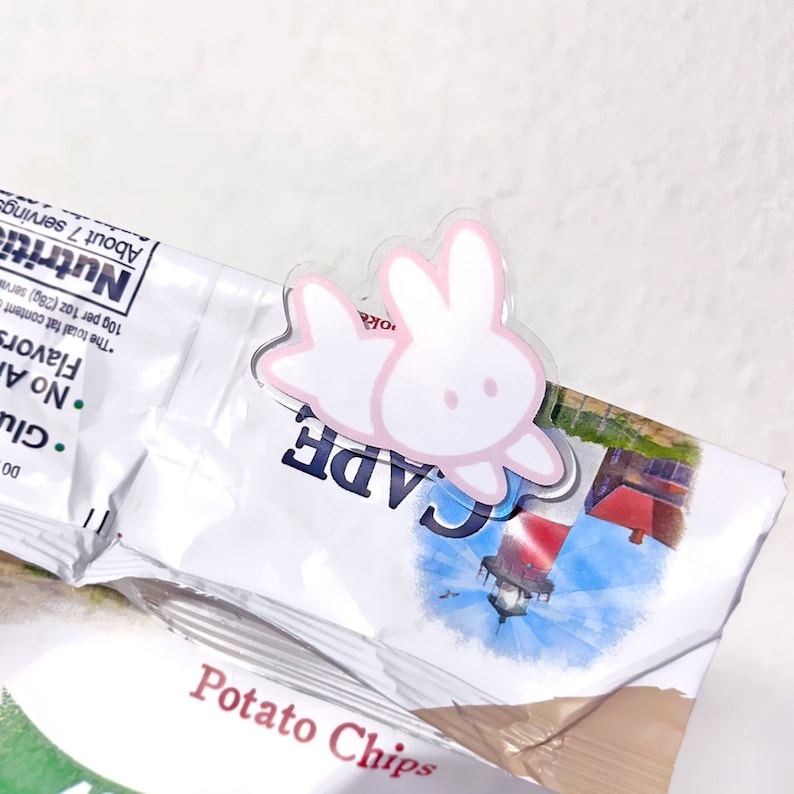 Op de afbeelding: Een transparante plastic chipclip met een roze konijnenontwerp. Het konijn is wit met roze oren en een roze omtrek. De clip is bevestigd aan een zak chips.