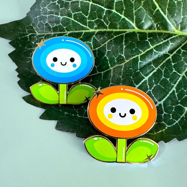May include: Two enamel pins shaped like flowers with smiling faces. The blue flower has a gold star above it and the orange flower has a gold star to the left of it. Both flowers have green leaves.