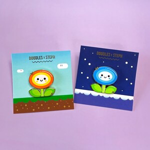 May include: Two enamel pins featuring cartoon flower designs. The first pin is a yellow and orange flower with a happy face and a star on top. The second pin is a blue flower with a happy face and a star on top. Both pins are on a white background with text that reads "DOODLES x STEPH".